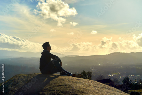 young boy sitting on top of a mountain looking to the horizon. enterprising man sitting on a rock with background sunset landscape. dark teenager with Latino features on a mountain