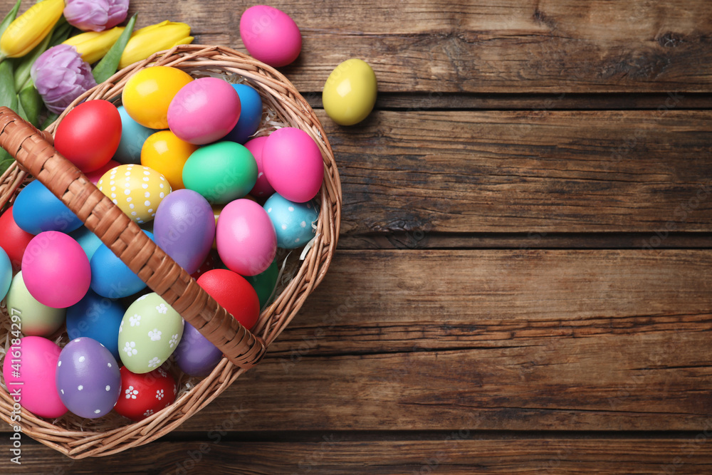 Colorful Easter eggs in wicker basket and tulips on wooden table, flat lay. Space for text Colorful Easter eggs in wicker basket and tulips on wooden table, flat lay. Space for text