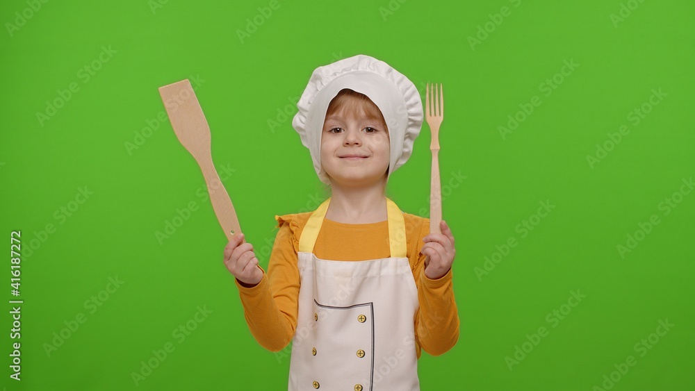 Fototapeta premium Child girl kid dressed as professional cook chef showing wooden fork and spatula, smiling, nods head in agreement on chroma key background. Nutrition, cooking school, education, food. Fun and humor