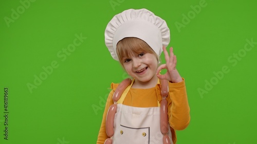 Charming child girl dressed in apron and hat like chef cook showing approval ok sign, invites you to come on chroma key background. Nutrition, family cooking school, children education, restaurant