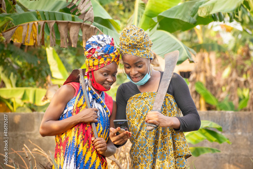 Two beautiful African female farmers with nose mask, looking happily into a smartphone and holding hoe and cutlass in a banana plantation 