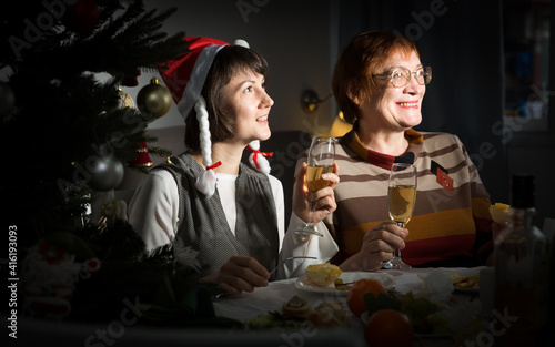 Cheerful mature woman celebrating New Year at home with her adult daughter  watching festive tv program together