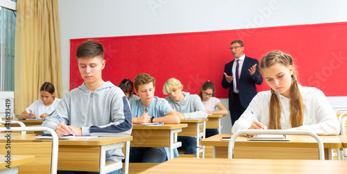 Smart teenagers studying in classroom, listening to lecturer and writing in notebooks