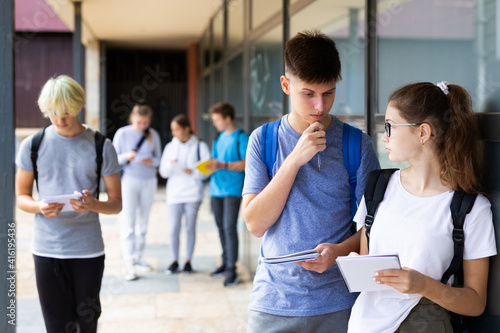 Two teenage students discussing while standing with workbooks near college building on autumn day.