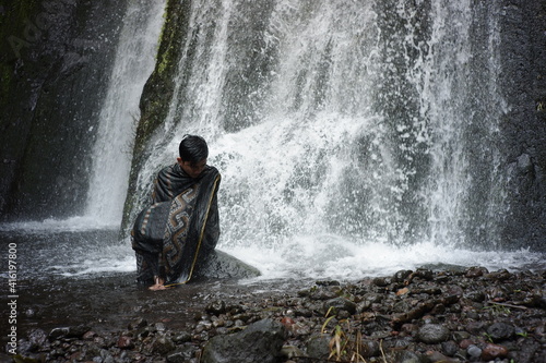 A portrait of an Asian man wearing a Lombok-Indonesian songket cloth enjoying the freshness of a waterfall in the middle of the forest. a traveler relaxing and bathing under a swift waterfall