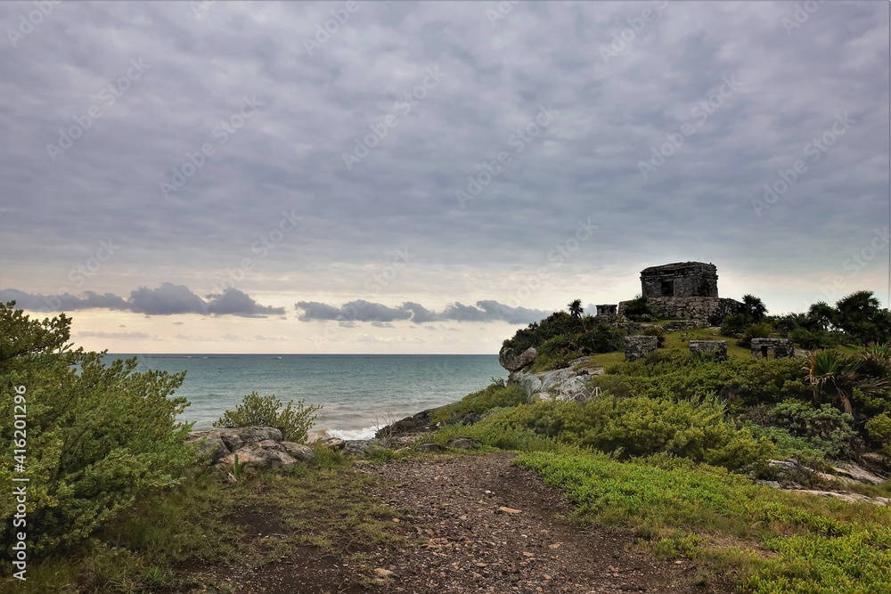 Ruins of the ancient Mayan city of Tulum on the shores of the Atlantic Ocean. A dilapidated building stands on a hill. Nearby are tropical vegetation and a path. There are clouds in the sky. Mexico