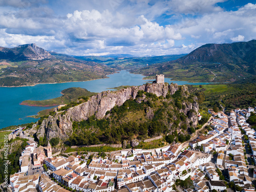 Aerial view of fortified castle on top of crag and white residential houses at bottom of hill in Zahara de la Sierra on background of turquoise lake, Spain
