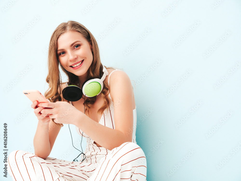 Naklejka premium Beautiful smiling woman dressed in summer clothes. Sexy carefree model listening music in headphones. Adorable and positive female posing near light blue wall in studio. Using her smartphone