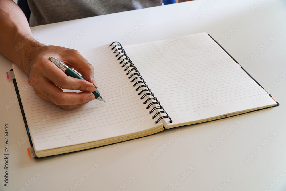 man's hand written in a spiral notepad placed on a wooden table with items. man's hand written in a spiral notepad placed on a wooden table with items.