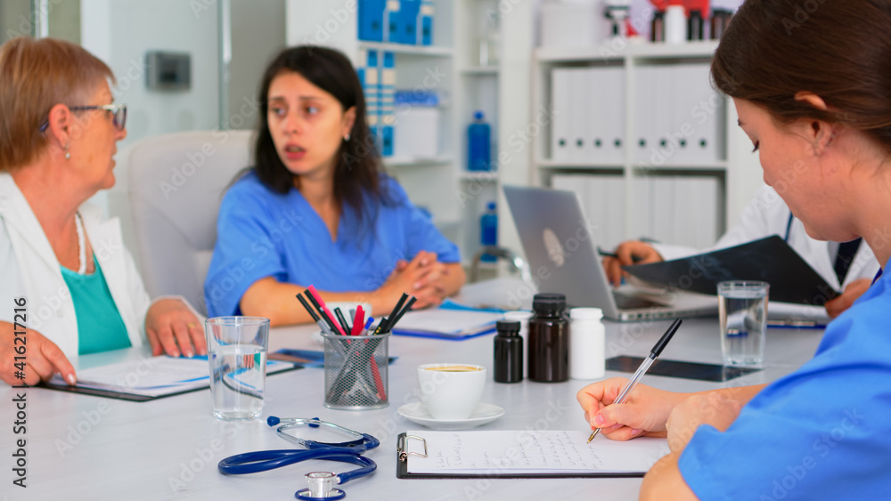 Obraz premium Nurse writing on clipboard while profesional teamworkers having medical meeting discussing in background in brainstorming office. Profesional doctors examining the symptoms of patient in meeting room.