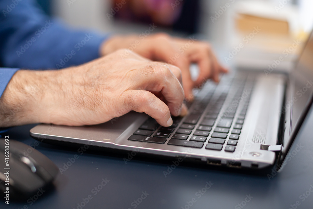 Close up of senior man hands typing on laptop keyboard. Elderly man entrepreneur in home workplace using portable computer sitting at desk while wife is holding tv remote Close up of senior man hands typing on laptop keyboard. Elderly man entrepreneur in home workplace using portable computer sitting at desk while wife is holding tv remote