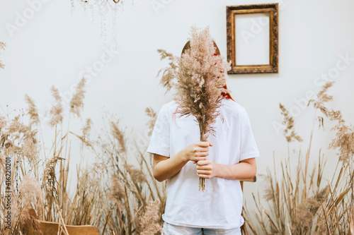 red haired girl closes her face with bouquet wearing a white shirt and denim shorts in a space decorated with dry grass and flowers in a retro style