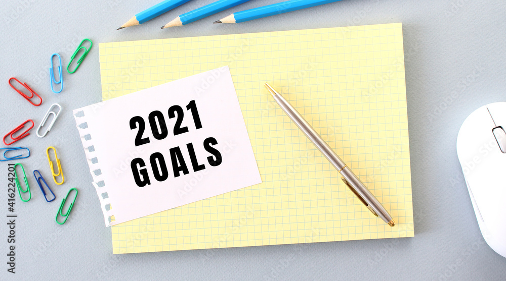 2021 GOALS is written on a piece of paper that lies on a notebook next to office supplies. 2021 GOALS is written on a piece of paper that lies on a notebook next to office supplies.