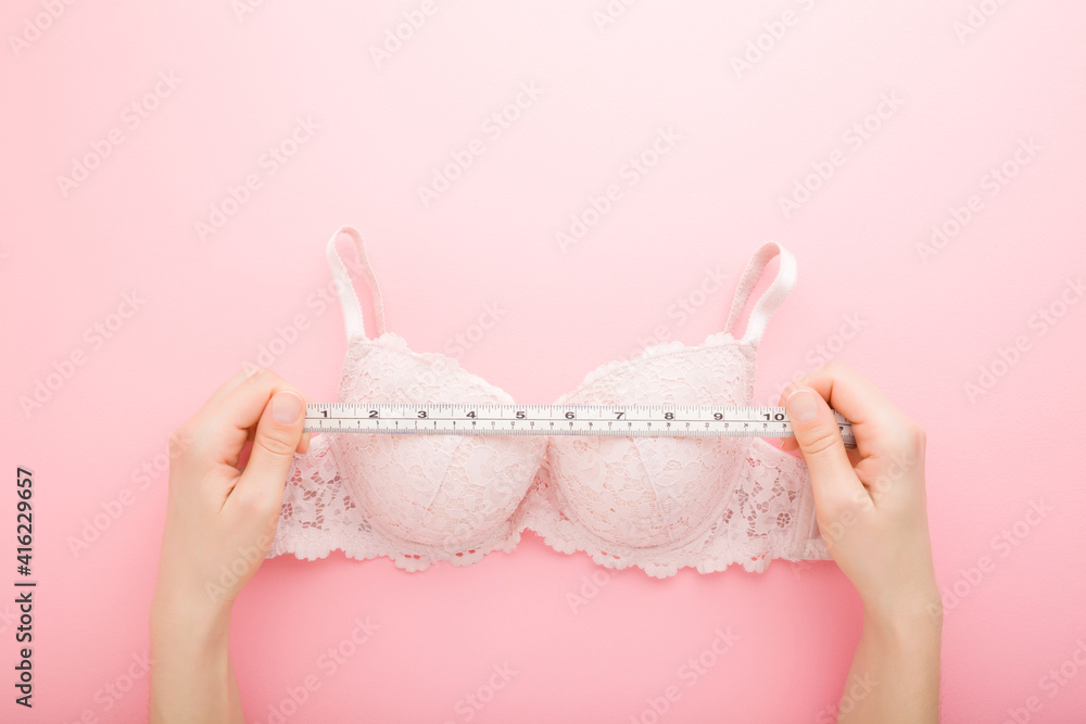 Young adult woman hands holding white measure tape and measuring bra on light pink table background. Pastel color. Closeup. Point of view shot. Choosing right size. Top down view. - obrazy, fototapety, plakaty 