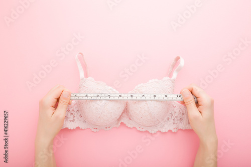 Young adult woman hands holding white measure tape and measuring bra on light pink table background. Pastel color. Closeup. Point of view shot. Choosing right size. Top down view.