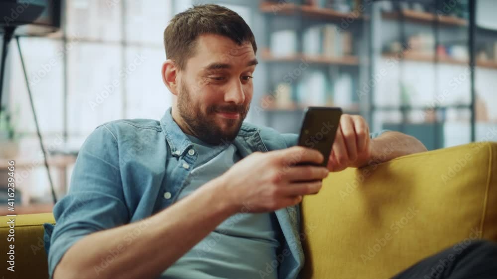 Happy Handsome Caucasian Man Using Smartphone in Cozy Living Room at Home. Man Resting on Comfortable Sofa. He's Browsing the Internet and Checking Videos on Social Networks and Having Fun. Happy Handsome Caucasian Man Using Smartphone in Cozy Living Room at Home. Man Resting on Comfortable Sofa. He's Browsing the Internet and Checking Videos on Social Networks and Having Fun.