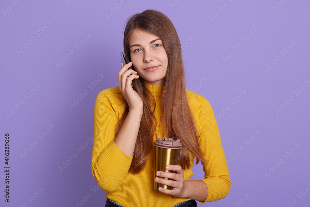 Positive female talks with interest and joy via cellular, wears casual attire, enjoys drinking aromatic coffee fro thermo mug, isolated over lila background, lady looks at camera. Positive female talks with interest and joy via cellular, wears casual attire, enjoys drinking aromatic coffee fro thermo mug, isolated over lila background, lady looks at camera.