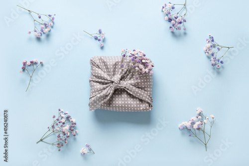 Fototapeta Naklejka Na Ścianę i Meble -  Spring eco-friendly gift wrapped in textile with fresh flowers on blue background. Sustainable gift. Traditional Japanese Furoshiki style. View from above.