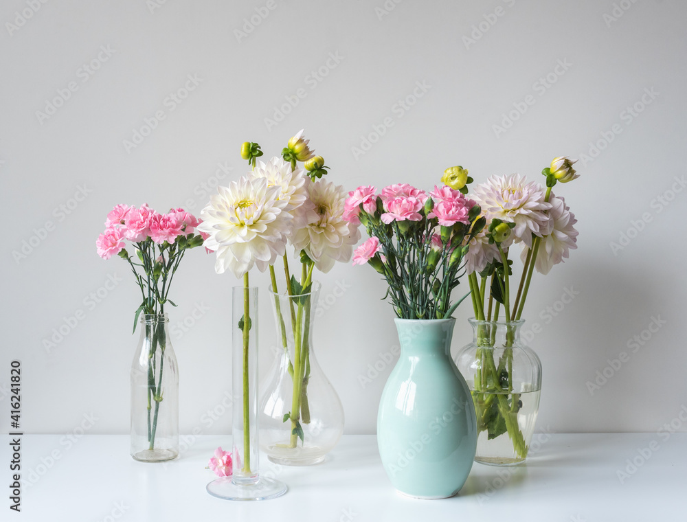 Close up of pink carnations and white dahlia flowers in glass and ceramic vases on white shelf against wall (selective focus)