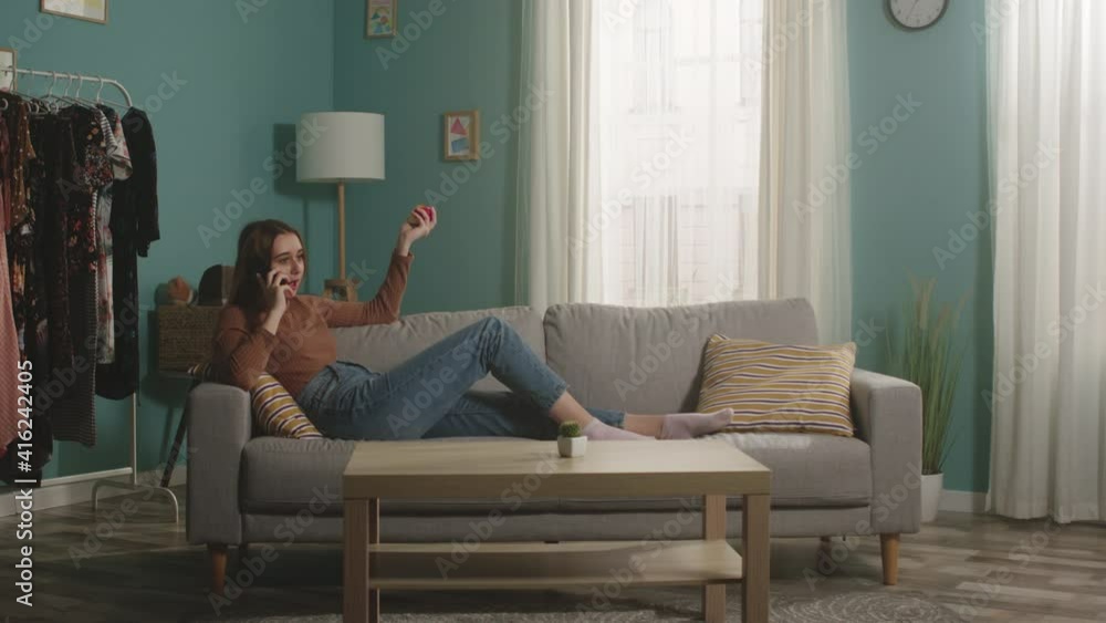 A young woman in blue jeans and a brown sweater lies on a beige sofa on the background of the window, holding a red apple and talking on the phone. General plan. A young woman in blue jeans and a brown sweater lies on a beige sofa on the background of the window, holding a red apple and talking on the phone. General plan.