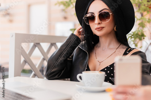 Fashionable woman in elegant hat in fashion sunglasses in trendy clothes sits and makes selfie on smartphone in cafe. Stylish girl in black youth wear photographs herself on summer poen terrace. photo