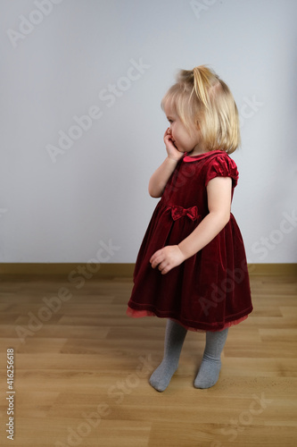 Wallpaper Mural little blonde girl 2 years old in a red velvet dress stands in profile in the room, is shy, holds her hands to her face, the concept of innocence, childhood Torontodigital.ca