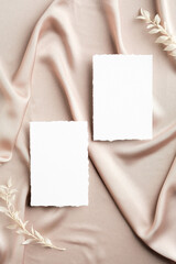 Flat lay wedding invitations mockups and flowers on beige silk. Top view with copy space. Elegant wedding stationery set.