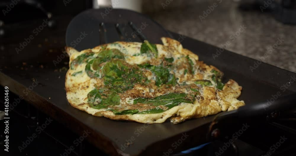 A chef slowly folds a spinach-filled egg omelet in half with a large spatula. A chef slowly folds a spinach-filled egg omelet in half with a large spatula.