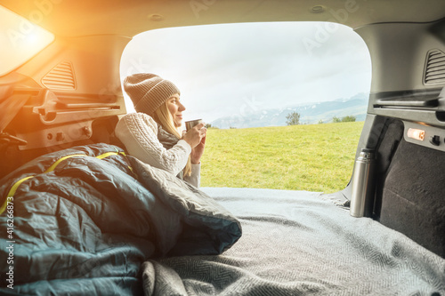 Girl resting in her car, journey made by car. Woman hiker, backpacker traveler camper in sleeping bag, relaxing, drinking hot tea on top of mountain. Road trip. Health care, authenticity,