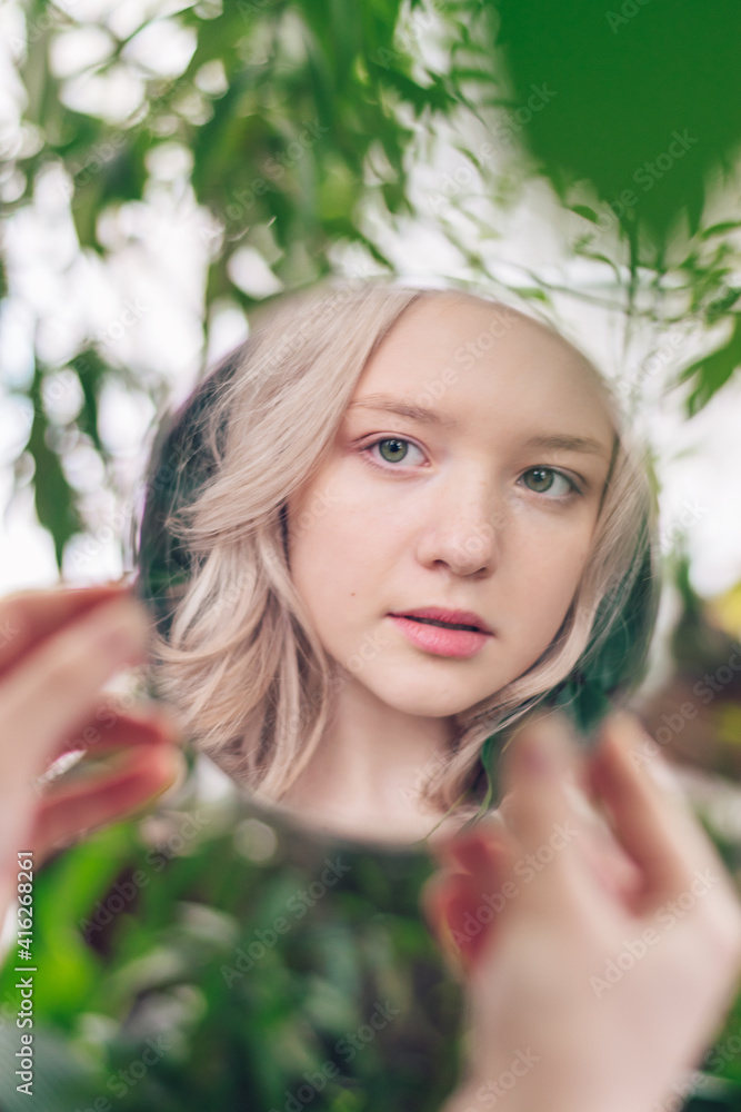 The reflection in the mirror of a beautiful blonde girl looking at herself. Plant leaves in the background. Psychological concept. Urban Jungle concept. The reflection in the mirror of a beautiful blonde girl looking at herself. Plant leaves in the background. Psychological concept. Urban Jungle concept.