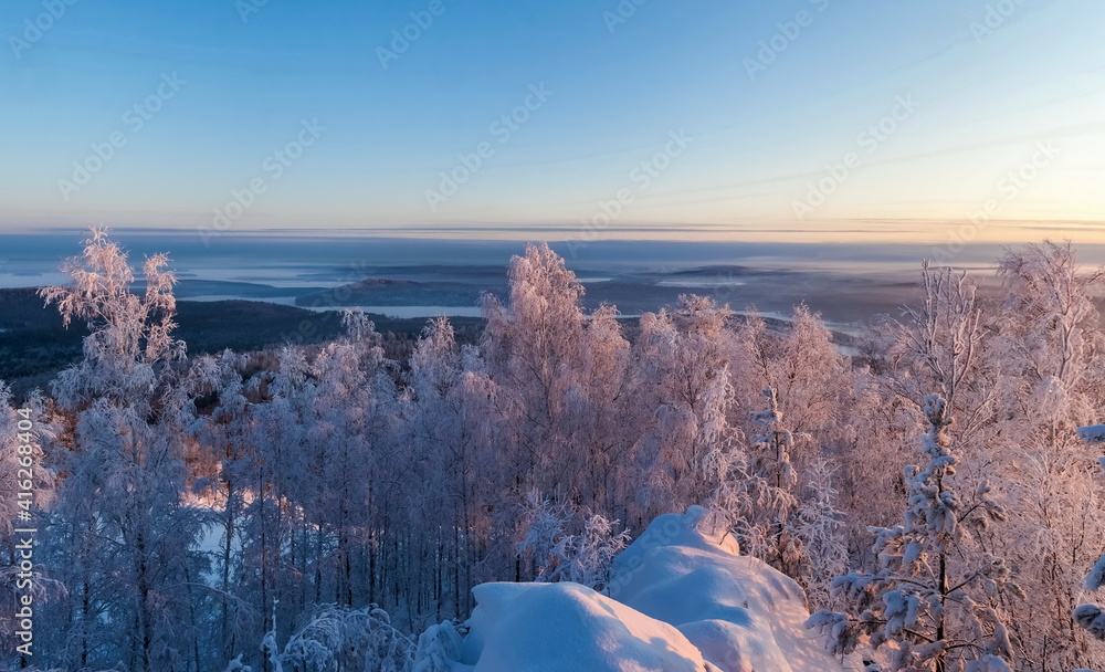 Winter landscape with snow-covered forest, mountains, sky, frosty haze in the rays of the setting sun