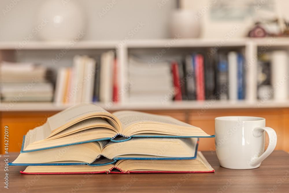 Open books, arranged one on top of the other, with a white cup of tea or coffee next to them, on a wooden surface, in front of the bookcase. Reading, studying, learning. Open books, arranged one on top of the other, with a white cup of tea or coffee next to them, on a wooden surface, in front of the bookcase. Reading, studying, learning.