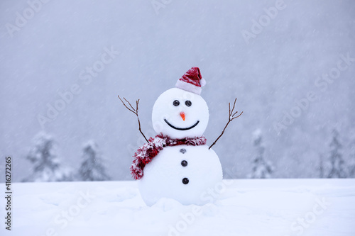 Snowman in santa hat and red tinsel on snowy field. Beautiful winter background. Merry christmas and happy new year. Cold foggy morning. Location place Carpathian, Ukraine, Europe. © Vitalii_Mamchuk