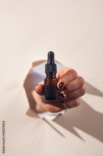 A woman's hand holds a bottle of face oil through a torn paper with hard shadows on the background. Concept of beauty.