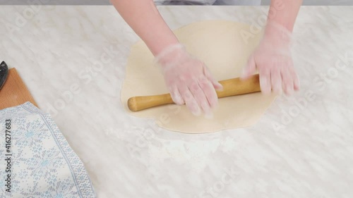 A woman with effort presses on a wooden dough roller, rolling out a thin dough on the table. Tight dough for dumplings. Close-up.