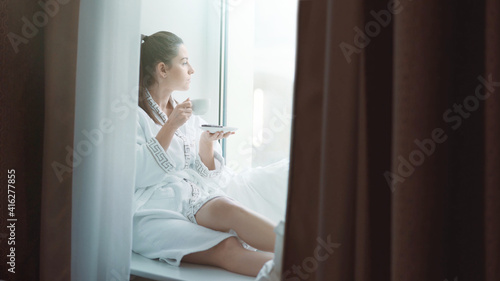 A young sweet brunette girl with brown hair in a white bathing bathrobe with patterns on a cloudy winter day off sits on the window sill, looks out the window and enjoys coffee or cocoa in a white cup