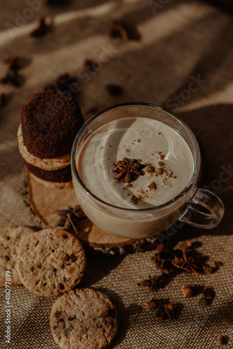 Beautiful creamy coffee in a cup with cookies and brownies carefully placed on the wooden surface with a brown background covered with beams of sunlight