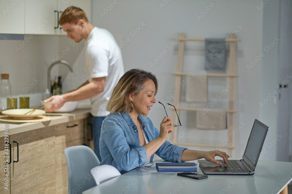 modern multinational couple at home in the kitchen. woman working or studying uses laptop. the husband in the background is cooking or cleaning. healthy relationships without gender stereotypes. modern multinational couple at home in the kitchen. woman working or studying uses laptop. the husband in the background is cooking or cleaning. healthy relationships without gender stereotypes.