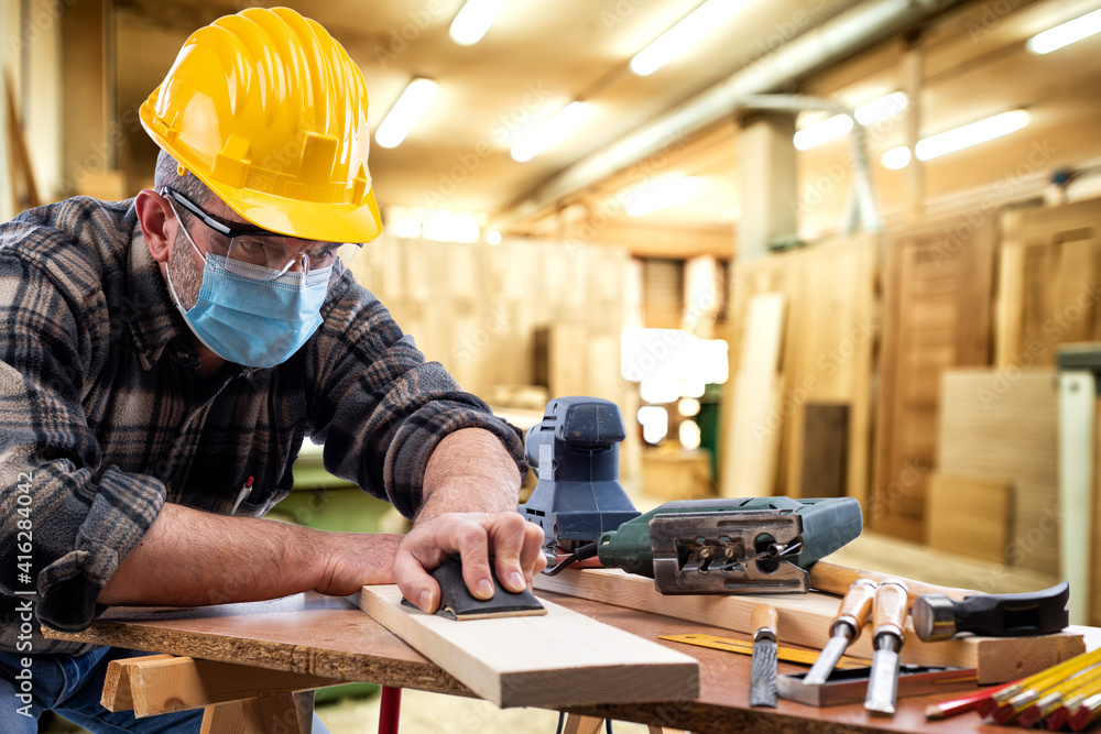 Carpenter worker at work in the carpentry workshop, wears helmet, goggles and surgical mask to prevent coronavirus infection. Preventing Pandemic Covid-19 at the workplace. Carpenter worker at work in the carpentry workshop, wears helmet, goggles and surgical mask to prevent coronavirus infection. Preventing Pandemic Covid-19 at the workplace.