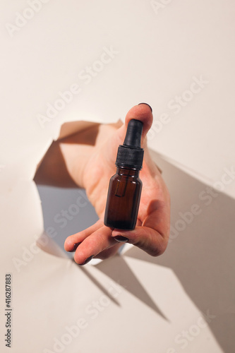 A woman s hand holds a bottle of face oil through a torn paper with hard shadows on the background. Concept of beauty.