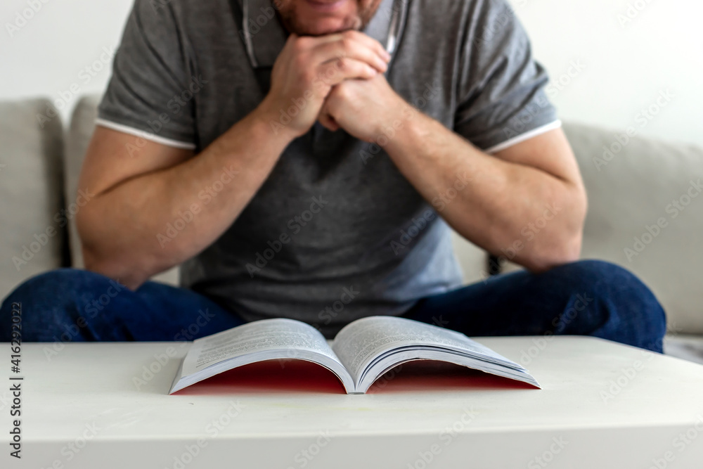 Selective focus of a young man sitting on sofa reading a book in a cozy loft apartment. Photo of a handsome bearded man in grey shirt sitting on the comfortable sofa, reading book and relaxing at home Selective focus of a young man sitting on sofa reading a book in a cozy loft apartment. Photo of a handsome bearded man in grey shirt sitting on the comfortable sofa, reading book and relaxing at home