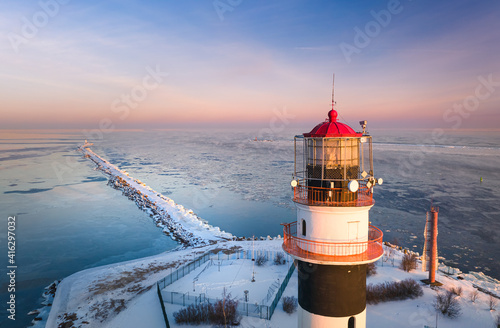 
Lighthouse at colorful sunrise at seaside in frosty winter morning. Snowy breakwater leading into frozen sea. 
