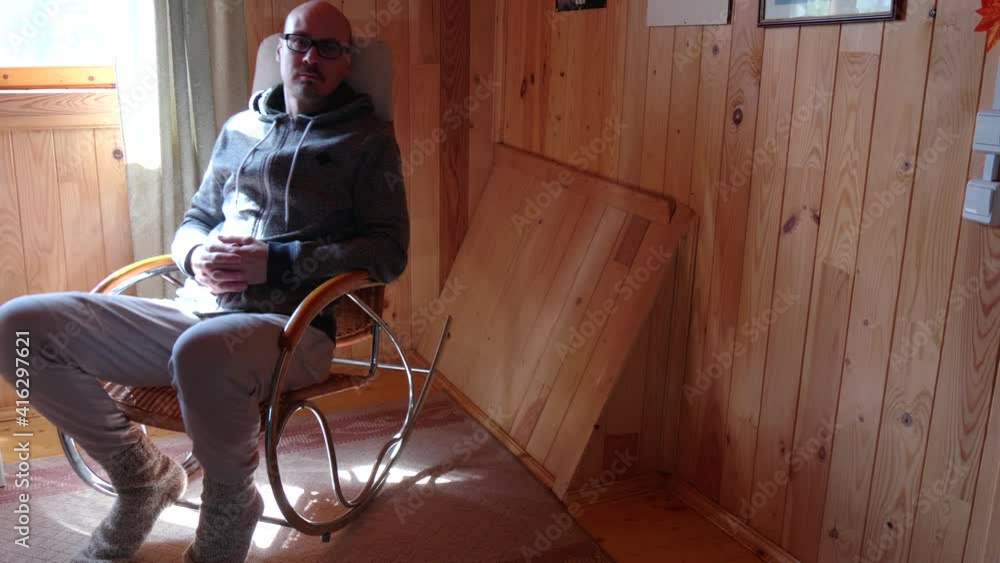 A young bald Caucasian man with glasses sways on wicker rocking chair ...