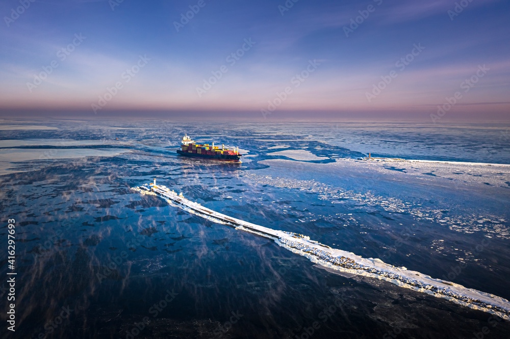 Cargo ship driving through frozen sea with colorful containers. Dark waters with ice blocks floating in sea.