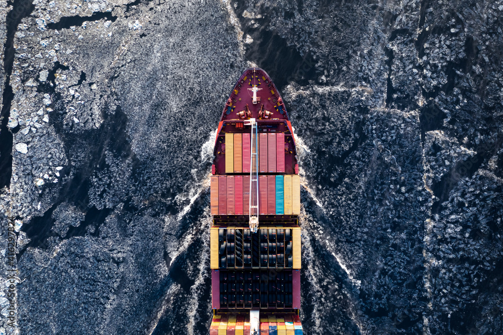 Cargo ship driving through frozen sea with colorful containers. Dark waters with ice blocks floating in sea.