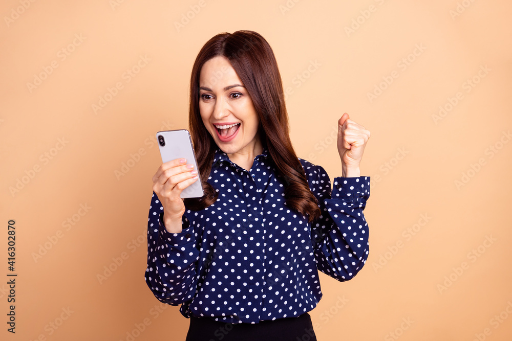 Photo of young excited woman happy positive smile look cellphone celebrate victory fists hands isolated over beige color background Photo of young excited woman happy positive smile look cellphone celebrate victory fists hands isolated over beige color background