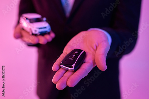 Cropped photo of a young attractive businessman in a black suit and white shirt holding a model car in the palm of his hand, isolated on a neon pink background. Car purchase, rental.