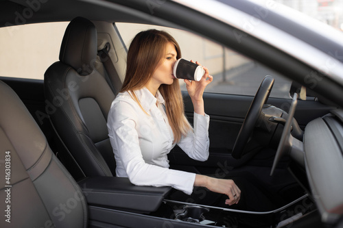 young pretty businesswoman drinking take away coffee, sitting inside of autonomous driverless electric car. self-driving vehicle. automotive technology.