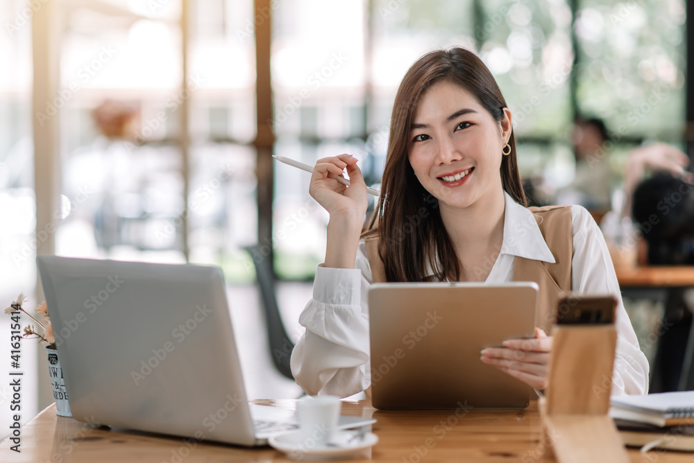 Charming Asian woman working on a laptop and tablet at the office. Looking at camera. Charming Asian woman working on a laptop and tablet at the office. Looking at camera.