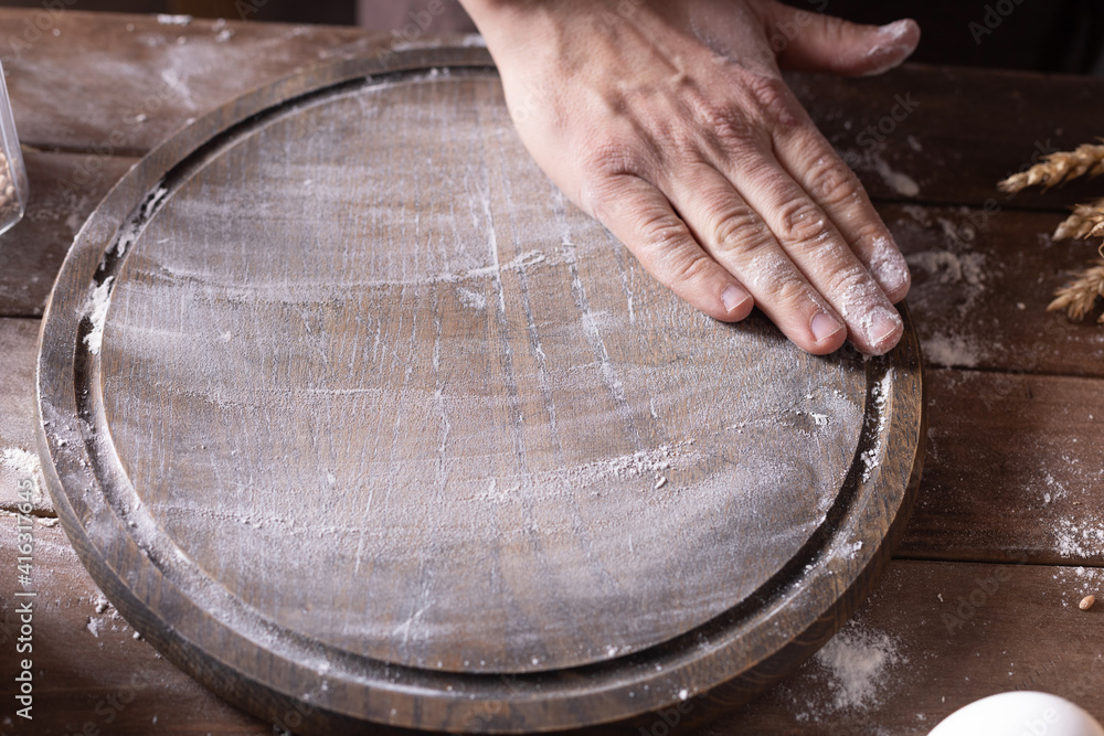 Baker male hand scattering flour for kneading dough at wooden pizza board
